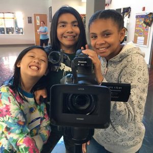 Three young future filmmakers holding a large camera grinning.