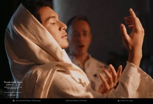 Person in robe gazing downward at their hand with person in background showing awed expression